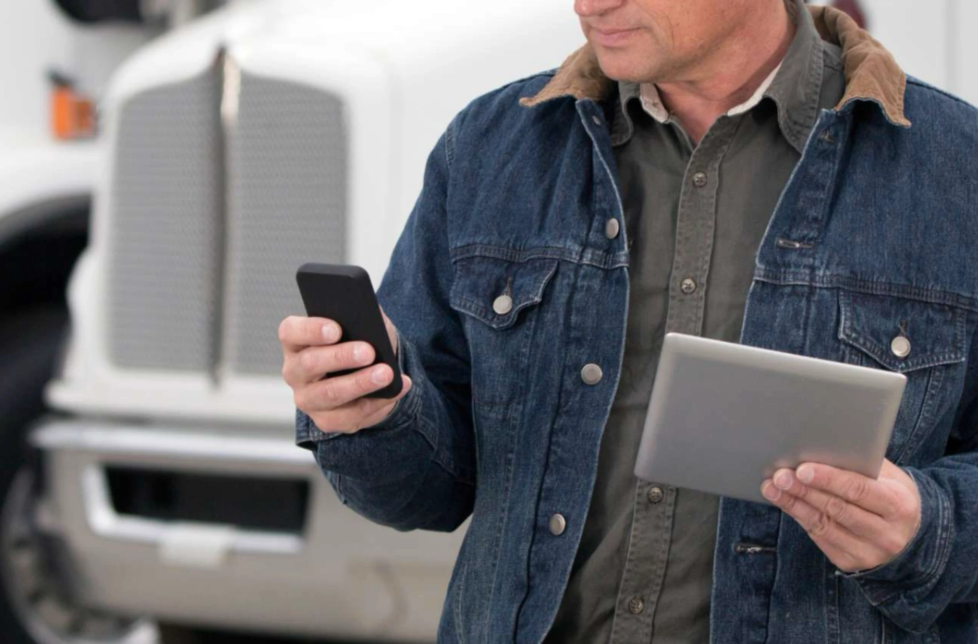 A man in a denim jacket checks a smartphone and holds a tablet, with the cab of a truck visible in the background.