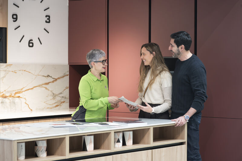 A Leroy Merlin specialist in a branded apron talking with a customer at a kitchen display counter, with product samples and catalogues visible.