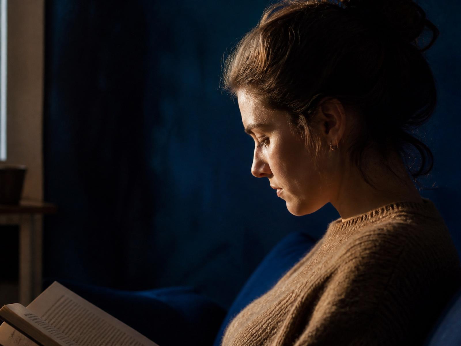 Side profile of a person reading quietly, warm window light on the face and deep indigo shadow behind.