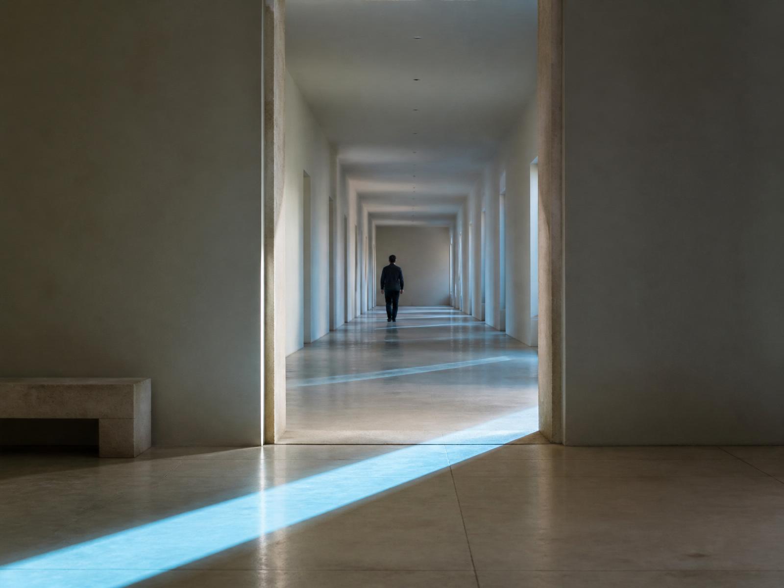 Long minimal stone corridor with a single figure walking through, a shaft of blue daylight crossing the floor between two thresholds.