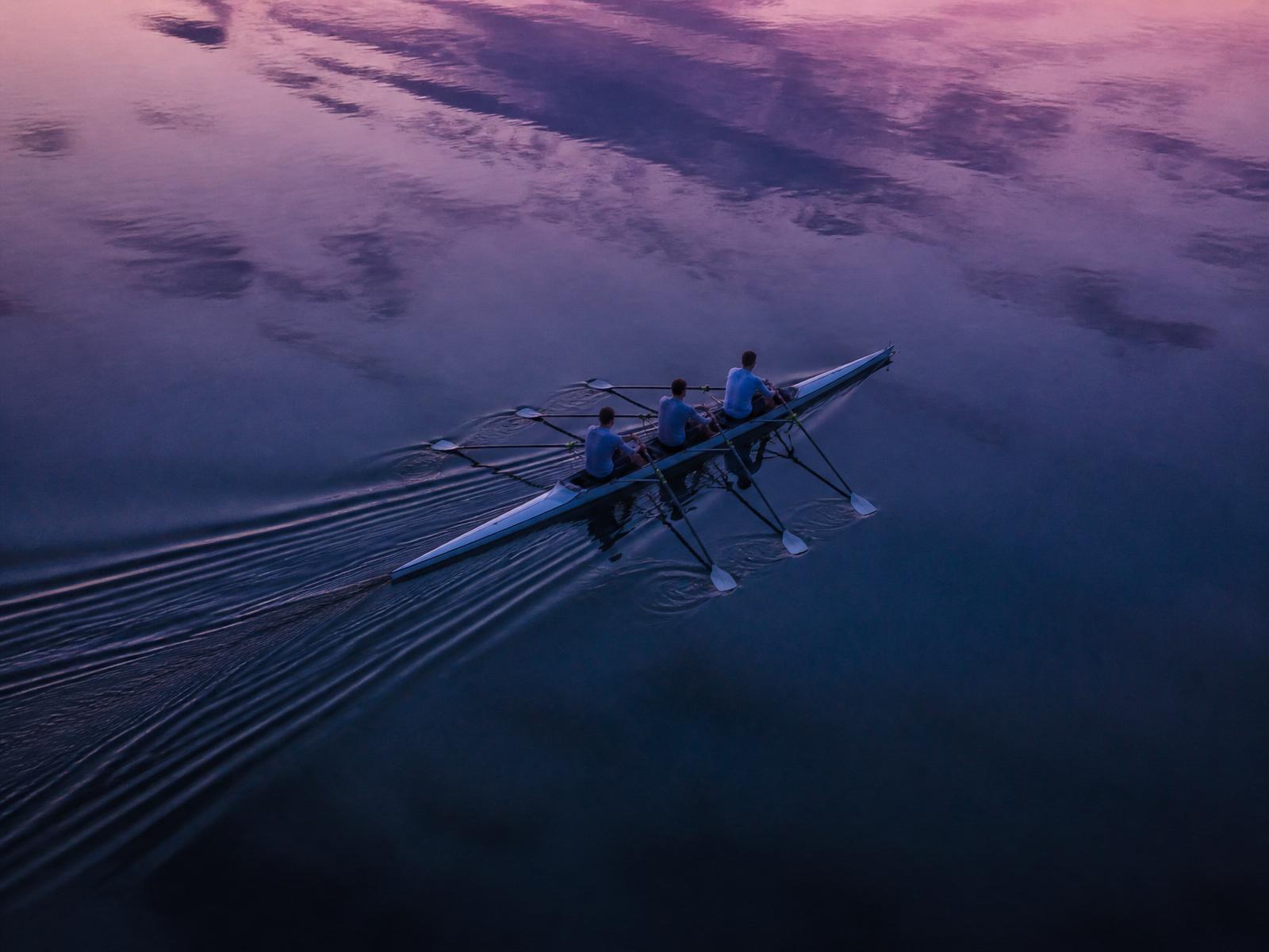 Aerial view of three rowers moving in synchrony across still water reflecting a violet and indigo dawn sky.