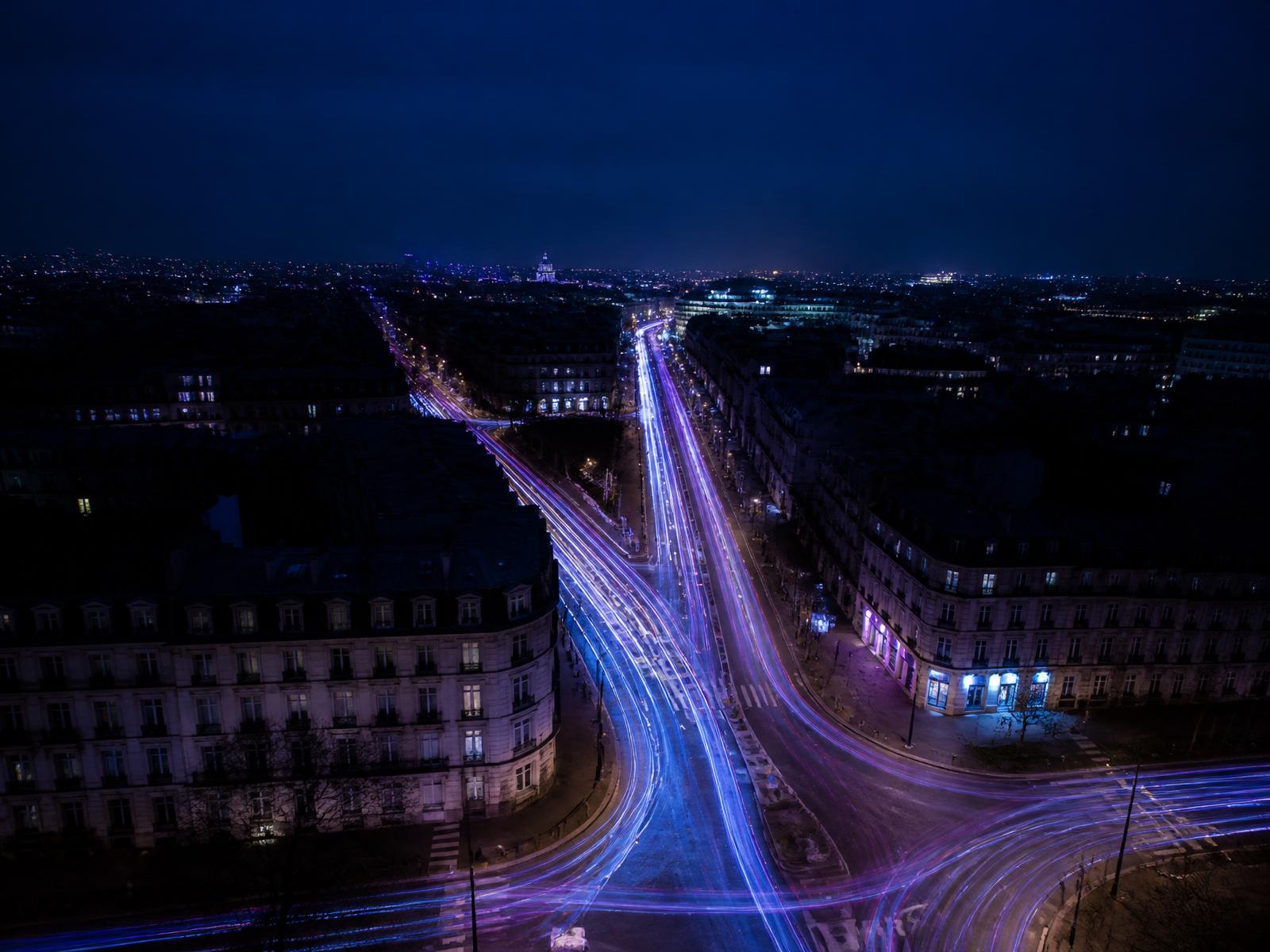 Aerial long exposure of a European city intersection at night, indigo and violet light trails converging from multiple directions.
